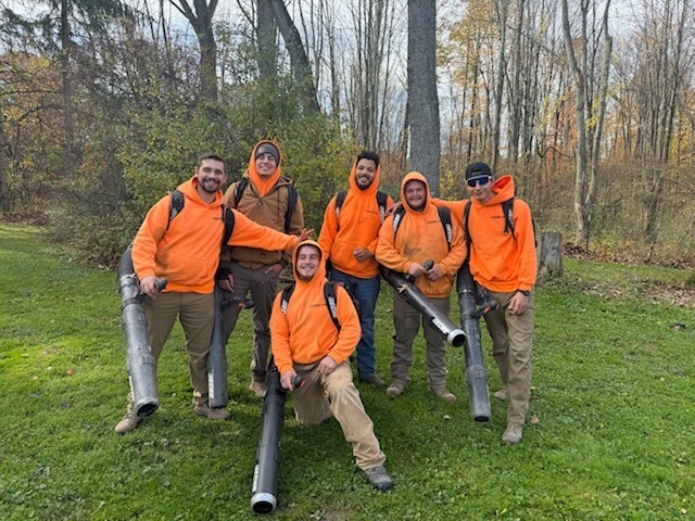 Six people in bright orange hoodies and work gear stand and kneel on a grassy area, each holding large leaf blowers. The background features leafless trees and bushes, indicating an autumn setting. They are smiling and appear to be enjoying their time outdoors.