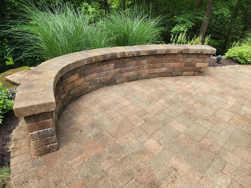 A curved brick bench surrounded by tall grass and greenery is set on a patterned brick patio. The background features lush trees, suggesting a serene outdoor setting.