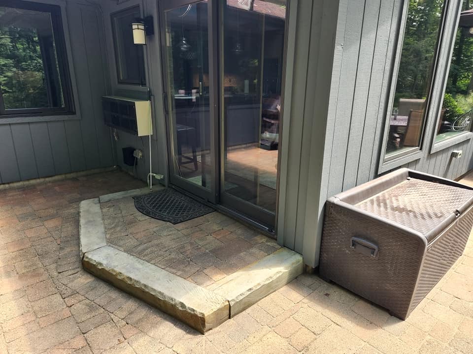 A patio with a brick floor leads to a sliding glass door entrance. There is a small raised concrete step with a welcome mat. An outdoor storage box is placed to the right of the door, and a wall-mounted air conditioning unit is visible on the left.