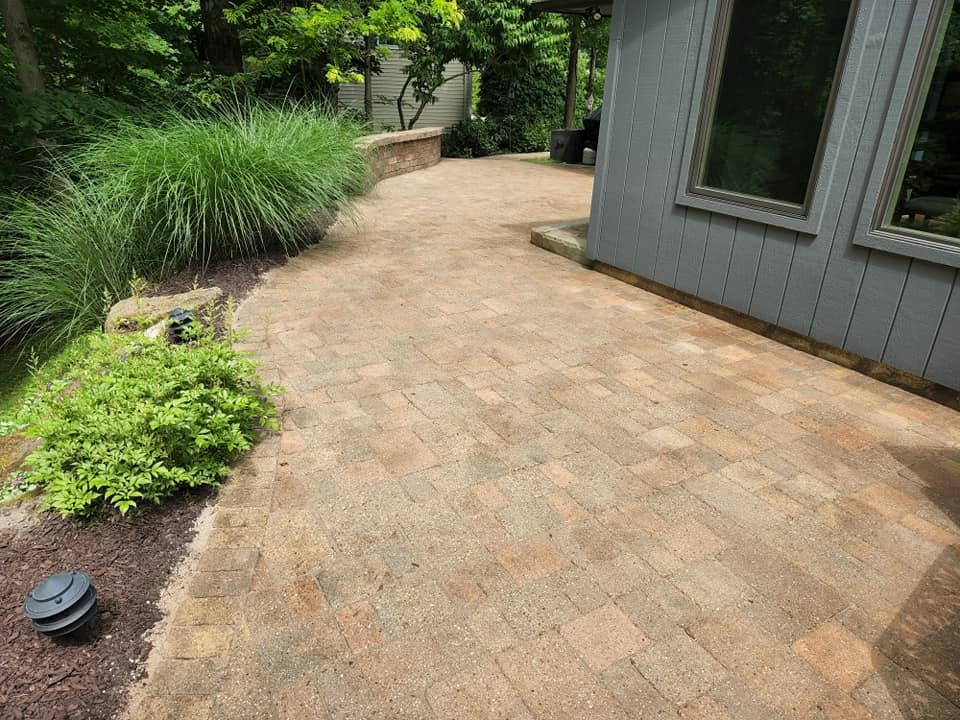 A paved brick patio extends alongside a gray building with large windows, bordered by lush green plants and trees. Theres a small garden light on the left and a raised stone garden bed in the background.