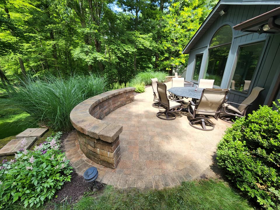 A sunny patio with stone flooring features a round table and swivel chairs. Theres a low stone wall with tall grass and vibrant greenery surrounding the area, leading to a peaceful forest view beside a house with large windows.