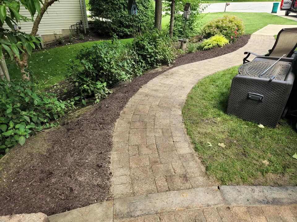 A curved brick pathway with mulch and green plants on the left side, and a lawn with patio furniture on the right. The path leads through a garden area, surrounded by trees and shrubs, with a concrete driveway visible at the end.