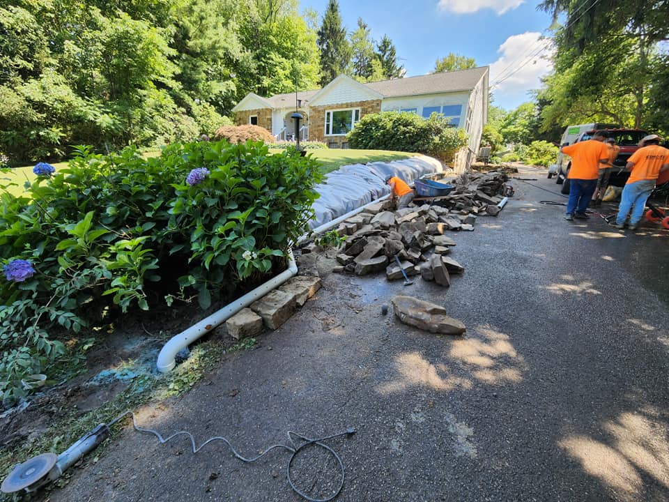 A team of workers in orange shirts repairs a driveway near a house. Rocks and equipment are scattered on the ground. Plastic sheets cover parts of the landscaping, and a hose is visible. Green shrubs and trees surround the area under a clear sky.