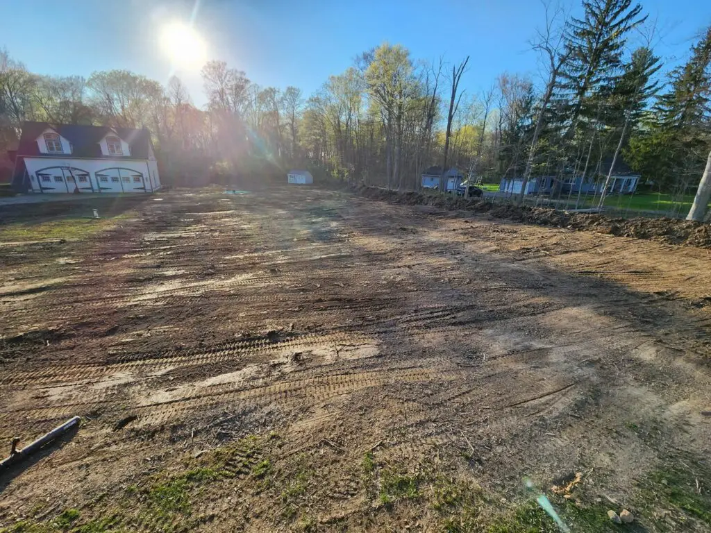 A cleared plot of land with visible tire tracks, set in a residential area. A house and a small shed are in the background surrounded by tall trees. The sun is shining brightly in the clear blue sky.