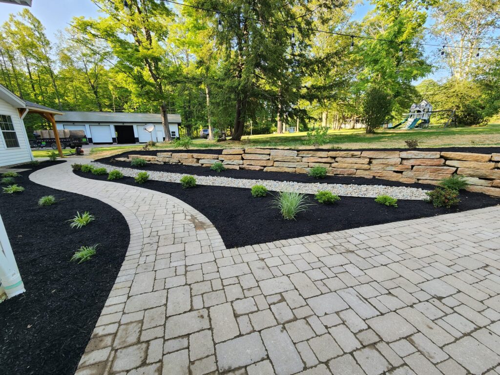 A backyard with a curved stone pathway leading past a garden bed with mulch, small plants, and a row of large stones in the background. A wooden playset is visible near lush green trees under a clear sky.