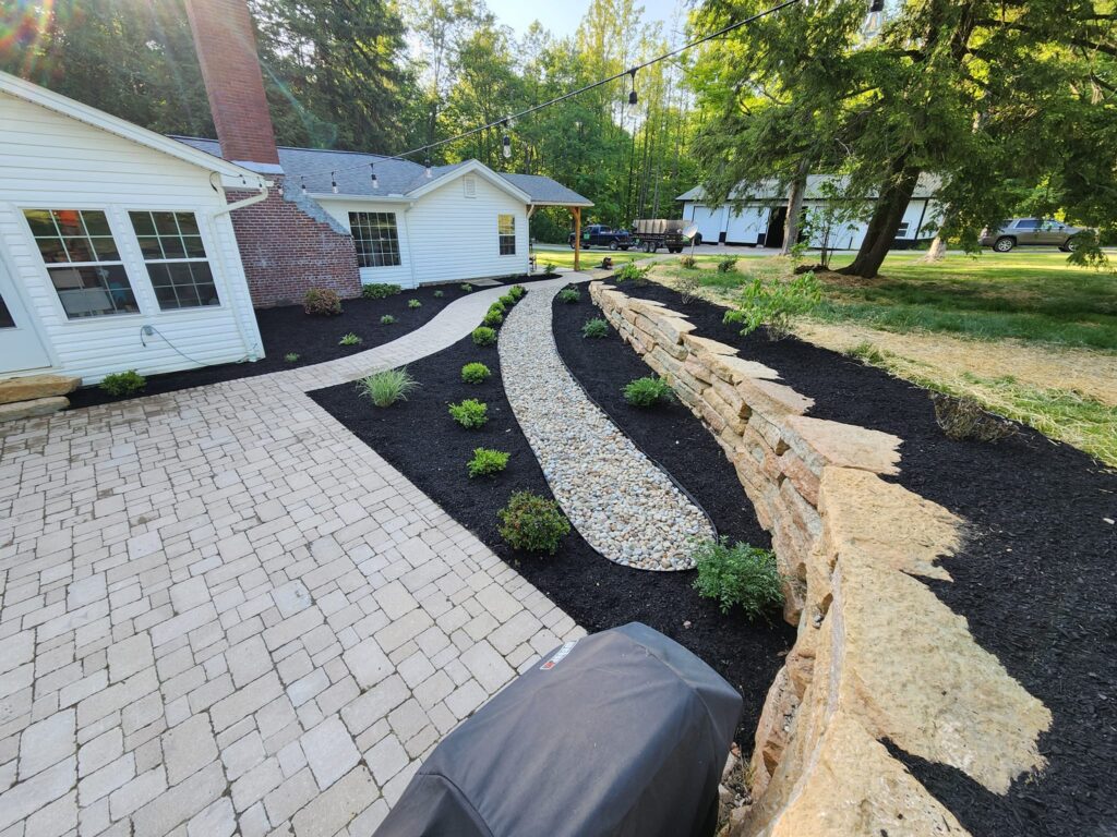 A landscaped yard with a stone pathway curves around a white house with a brick chimney. The path is bordered by black mulch and small green shrubs. A stone retaining wall runs parallel to the path, and a grill covers part of the patio.