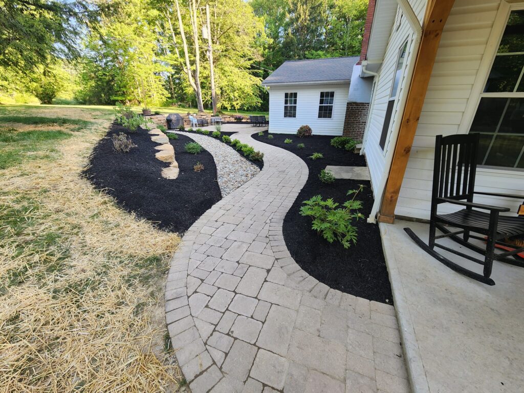 A light-colored, winding brick walkway leads alongside a house with a porch and rocking chair. Freshly mulched beds with young plants line the path, and green trees are visible in the background.