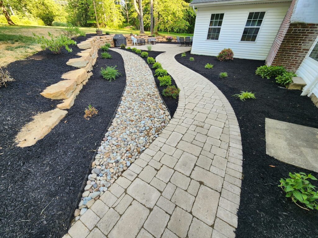 Curved stone pathway with adjoining pebble-filled section, bordered by black mulch and green plants, leading to a patio. The path runs alongside a house with a brick and white exterior. A seating area is visible in the background.