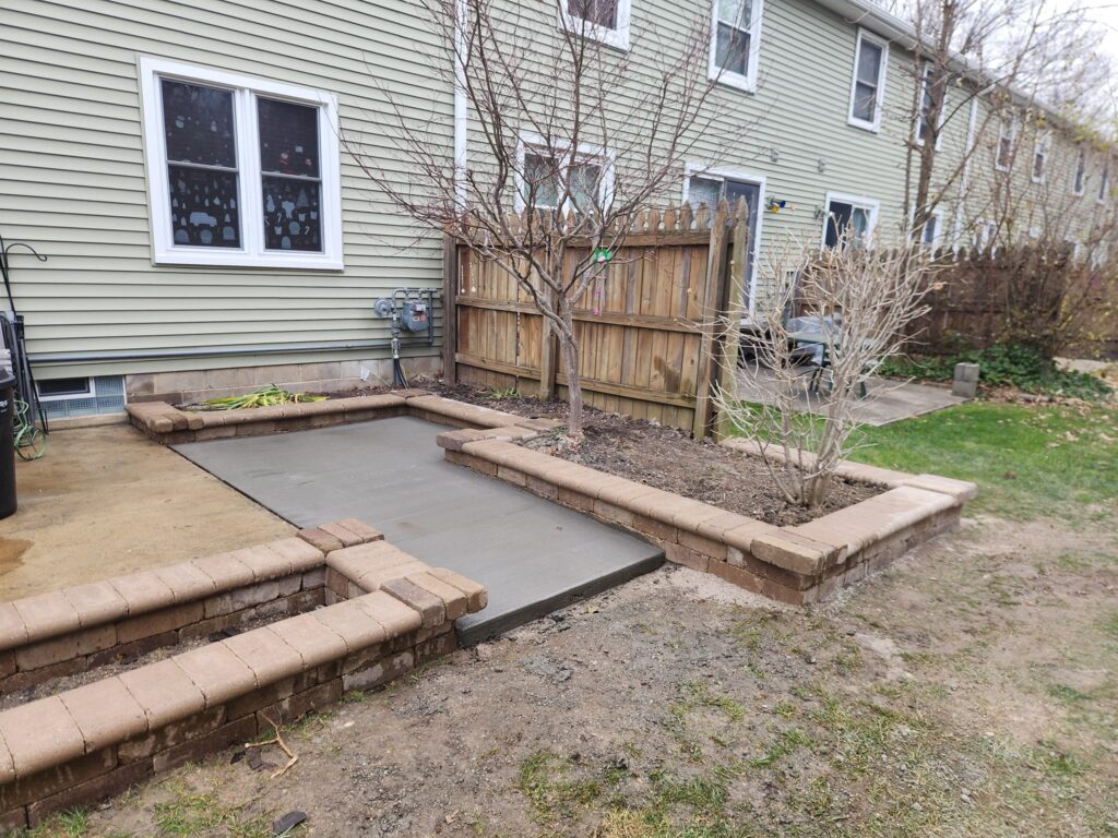 A backyard with a newly poured concrete patio surrounded by raised garden beds made of bricks. Bare tree branches are visible, and a wooden fence separates the yard from neighboring townhouses.