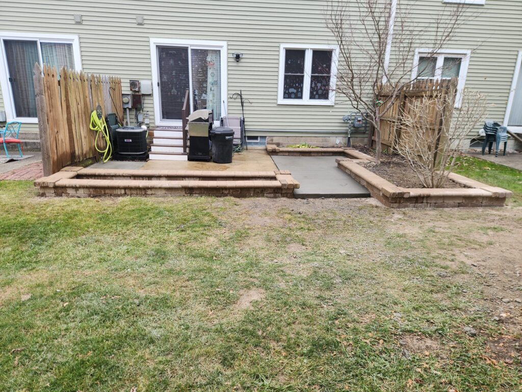 A backyard patio with a concrete and brick design. There are two black trash bins near the stairs leading to a house with glass doors. A garden hose hangs on a wooden fence, and a bare tree is within a raised brick flower bed. Grass covers part of the yard.