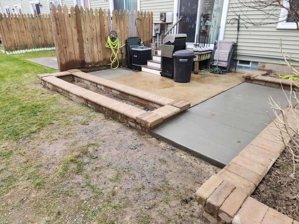 Backyard with new concrete steps and pathway. The area features a wooden fence, a garden hose reel, and a barbecue grill. The surrounding yard has grass and a patch of bare soil.