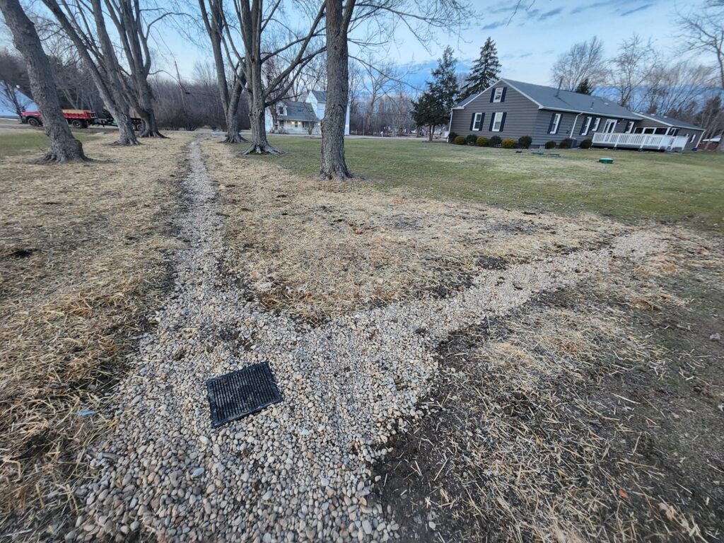 A gravel drainage path curves around bare trees on a grassy lawn. The path leads to a gray ranch-style house with white trim. The sky is cloudy, and the background features more trees and houses.