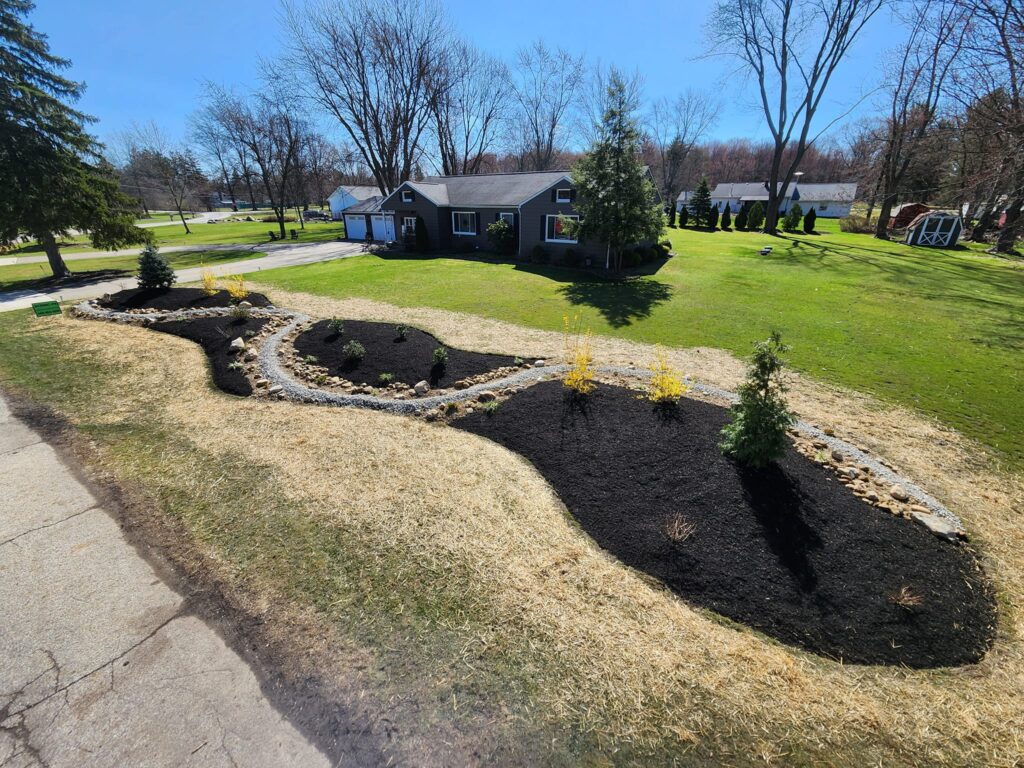 A landscaped front yard with a winding stone-bordered garden and freshly planted shrubs. The garden has dark mulch and is surrounded by a straw-covered area. A house with a gray exterior and large windows is in the background with bare trees.