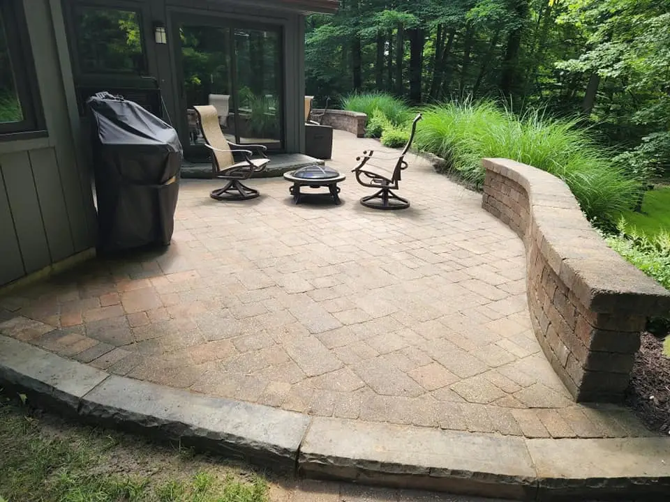 A paved patio features two swivel chairs around a fire pit, with a grill covered by a black cover nearby. A curved brick wall encloses the area, surrounded by tall grasses and trees in the background.