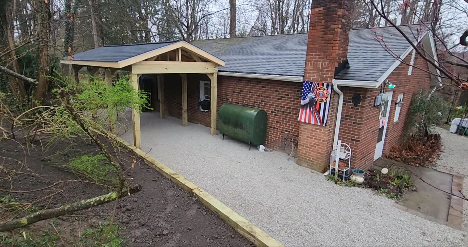 A brick house with a gray roof and a chimney displays an American flag. A green oil tank is beside the house. A new wooden porch structure extends over a gravel path bordering a landscaped yard with trees.