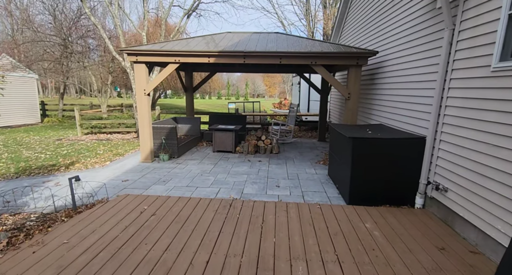 A backyard scene featuring a wooden gazebo with a metal roof on a stone patio. Inside, theres a fire pit with stacked logs and patio chairs. The area is surrounded by autumn trees and a wooden fence, adjacent to the side of a house.