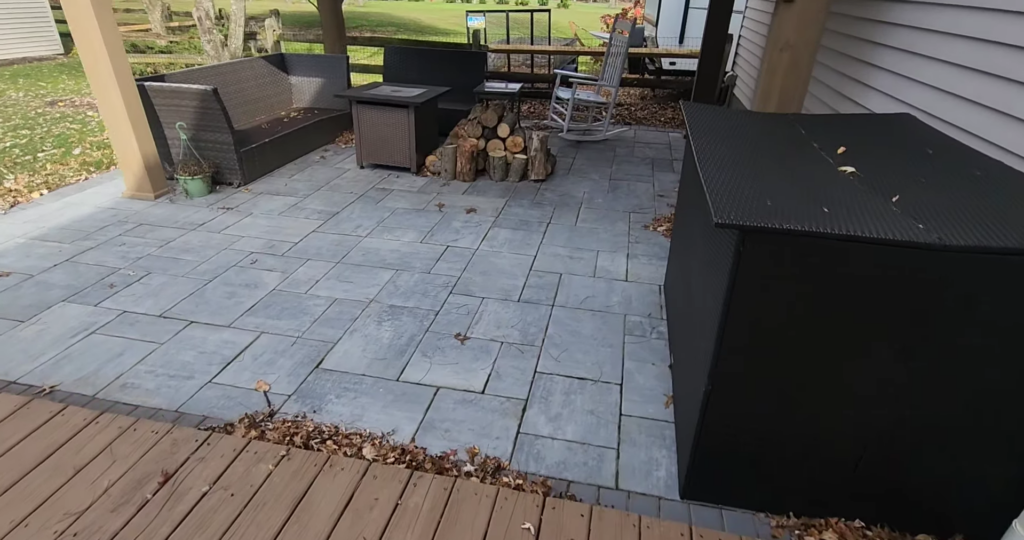 A patio with gray stone tiles features a wicker loveseat, a fire table, a stack of firewood, and a rocking chair. The patio is partially covered and bordered by a wooden deck. Fallen leaves are scattered on the ground.