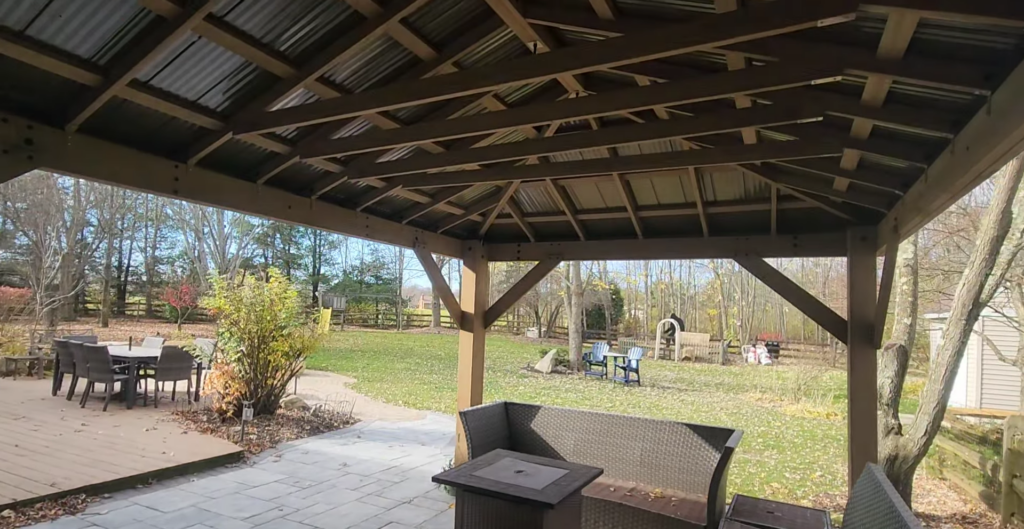 A backyard scene with a wooden gazebo covering a stone patio. There is a wicker sofa and a square fire pit. In the background, a table with chairs is on a wooden deck, and the yard is spacious with trees and scattered fallen leaves.