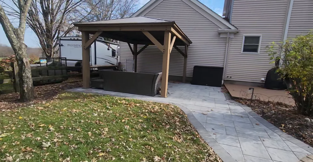 A backyard with a wooden gazebo over a seating area, surrounded by paved stones. Fallen leaves are scattered across the grass and pavement. A white trailer is parked nearby, and the house exterior is visible. Trees frame the space.