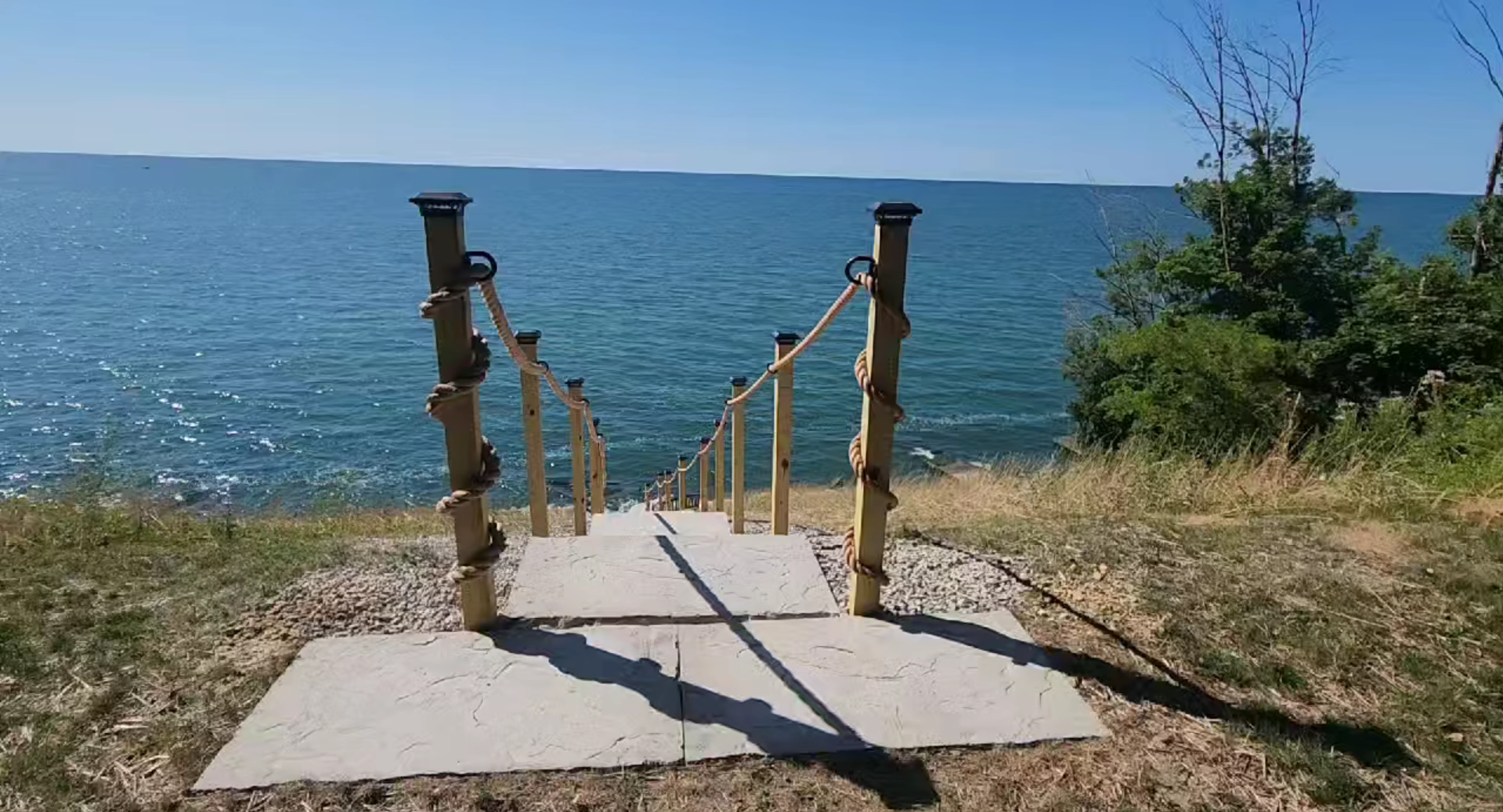 Wooden steps with rope handrails lead down to a rocky shoreline next to a large, calm body of water under a clear blue sky. Dense greenery is visible on the right side of the steps.