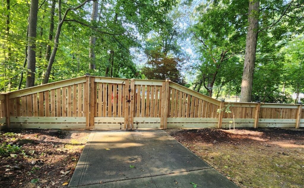 A wooden fence with a central gate runs along a paved path in a lush, green forested area. The fence has an angular design with tall posts and horizontal planks. Sunlight filters through the trees, casting dappled shadows on the ground.