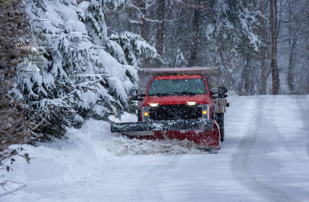 A red snowplow truck clears a snow-covered road surrounded by snow-laden pine trees. Snow is falling steadily in a wintry forest setting. The trucks headlights are on, illuminating its path through the thick snow.