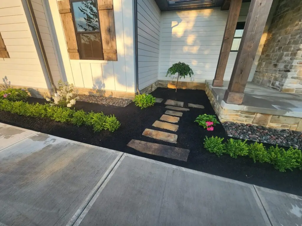 A landscaped front yard with a stone path leading to a house entrance. The garden features neatly trimmed green shrubs, a small tree, and bright pink flowers. The house has wooden shutters and a mix of stone and white siding exterior.