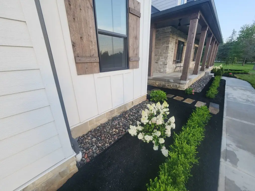 View of a modern house with white siding and rustic wooden shutters. The front features a stone porch with wooden pillars. A neatly landscaped garden includes white flowering plants and green shrubs along a walkway with stepping stones.
