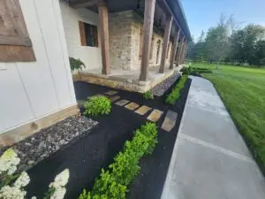 A landscaped garden with a stone pathway in front of a house with stone and wood accents. The garden features small shrubs, flower beds with mulch, and cobblestones, adjacent to a concrete walkway. A lawn and trees are in the background.