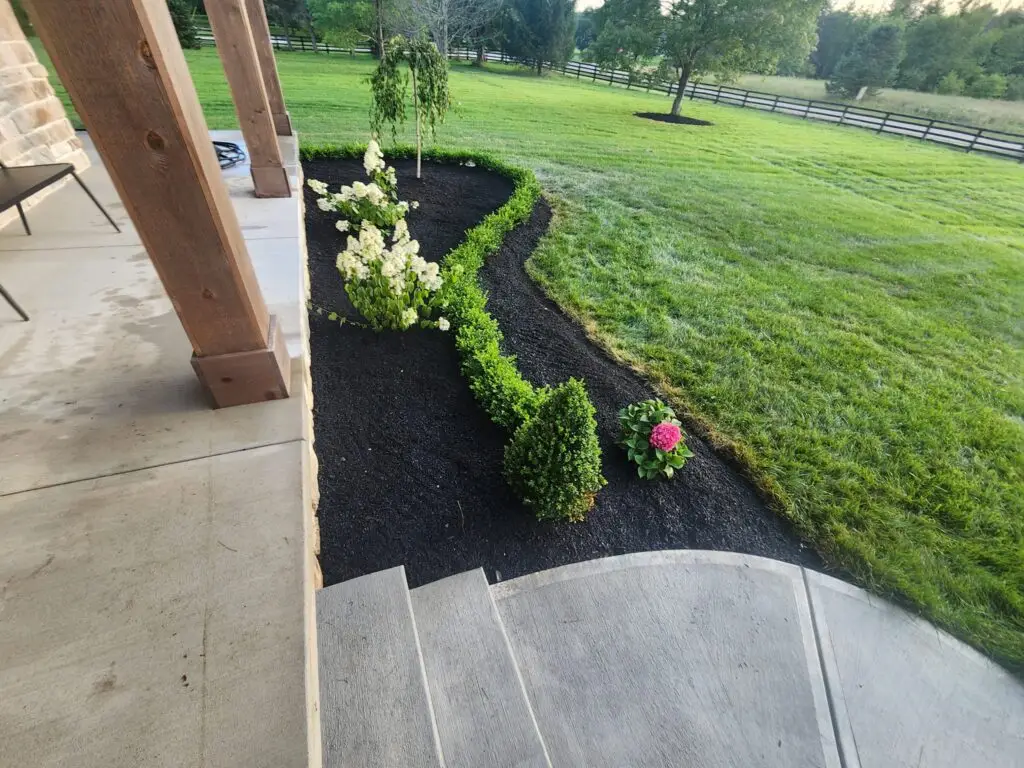 A landscaped garden with neatly trimmed bushes, flowering plants, and dark mulch lines a stone patio. Steps lead down to a lush green lawn surrounded by a wooden fence and trees in the background.
