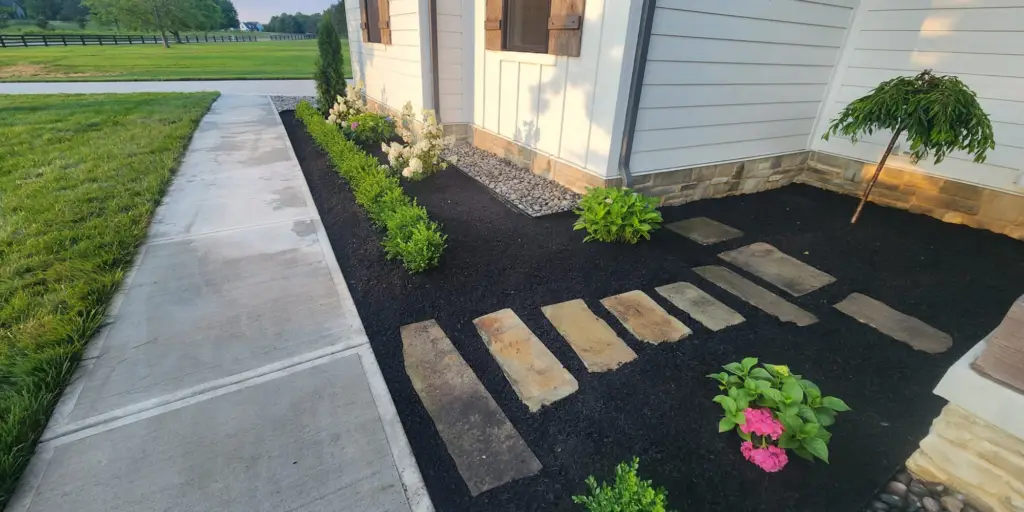 A landscaped garden with a stone walkway beside a house. The garden features black mulch, small shrubs, pink flowers, and a white flowering bush near the house. A concrete path runs alongside, bordered by a grassy lawn.