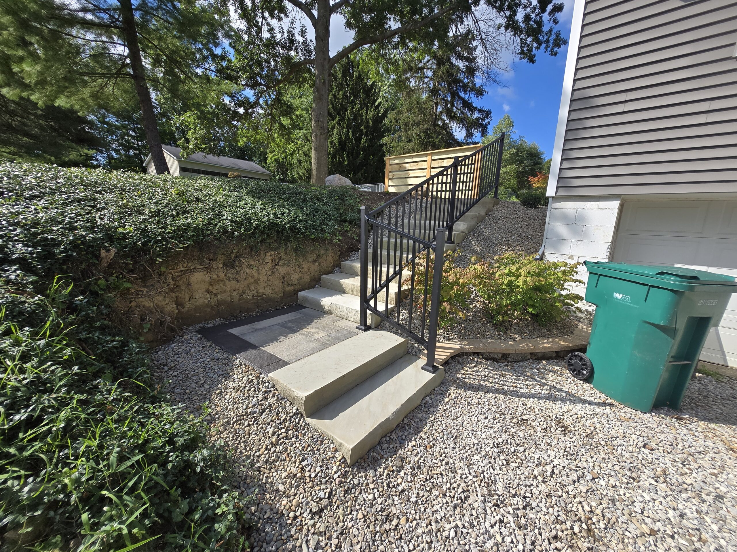 Concrete steps with a black metal railing lead up a small hill next to a house with gray siding, showcasing a Hardscape Transformation Ohio style. Gravel surrounds the area, with a green trash bin nearby and shrubs and trees in the background.
