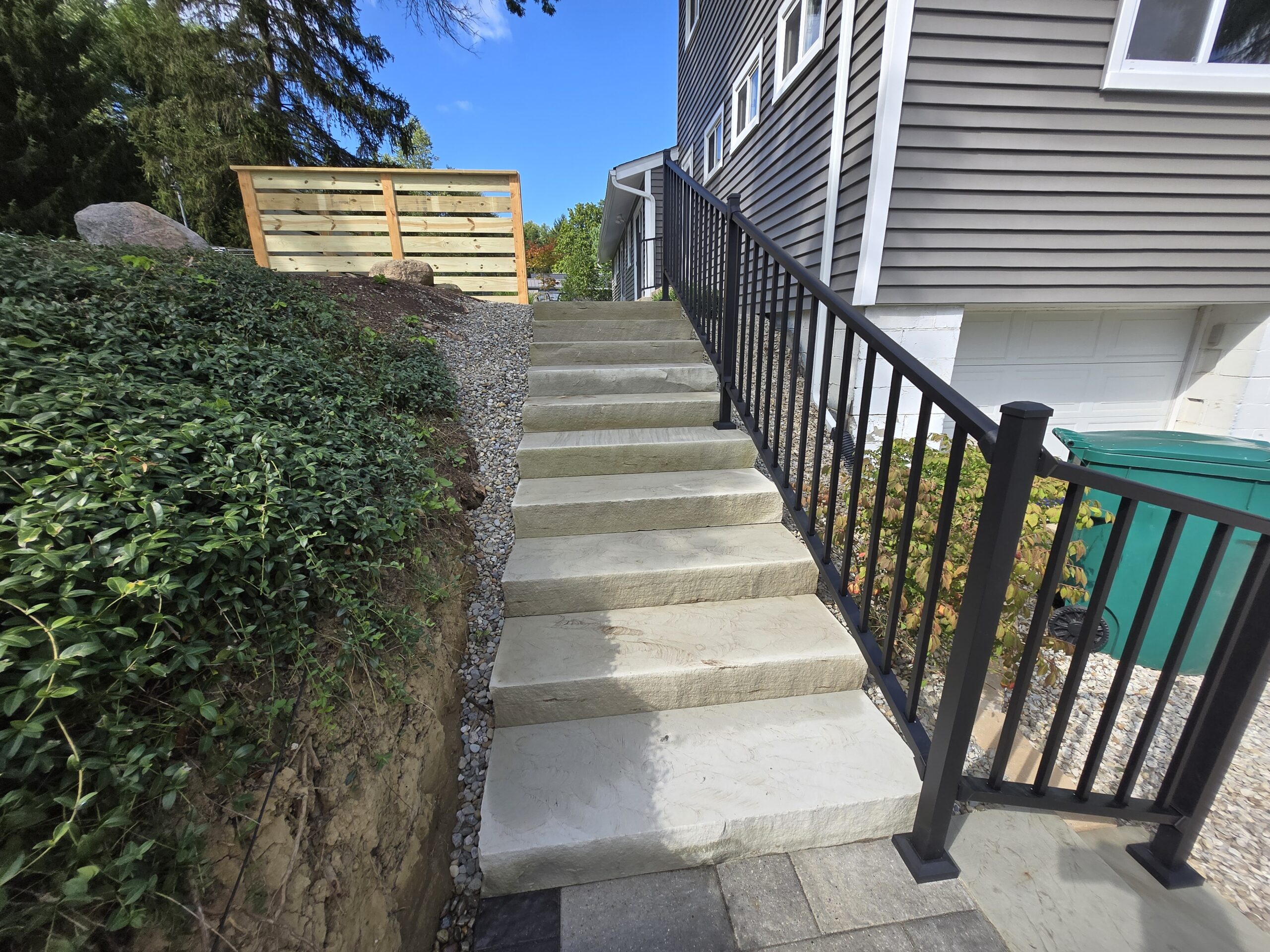 Stone steps with a black metal railing lead up beside a house with gray siding—a great example of Hardscape Transformation Ohio. A green garbage bin sits near the bottom, surrounded by rocks, greenery, and a wooden fence above.