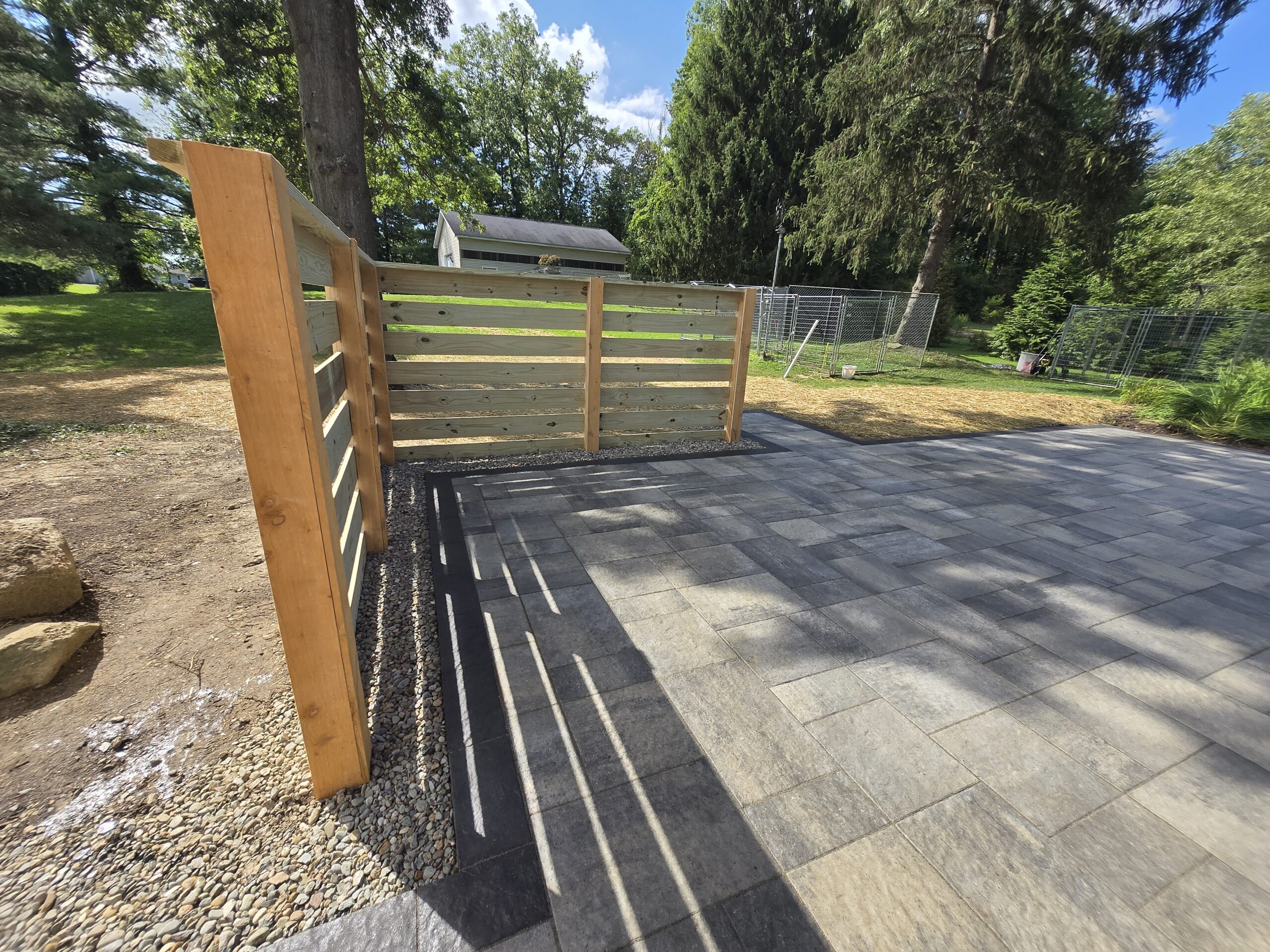 A newly constructed patio with gray stone tiles and a wooden privacy fence showcases a stunning hardscape transformation Ohio style, surrounded by grass, trees, and a small shed in the background on a sunny day.