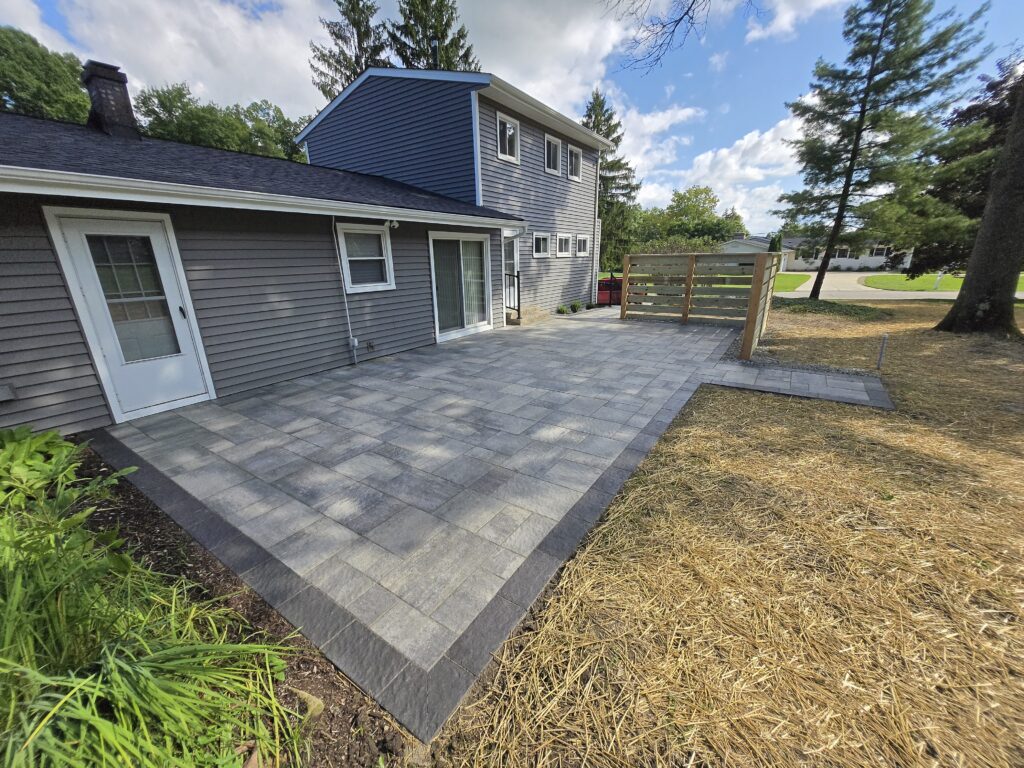 A modern backyard patio showcases a Hardscape Transformation Ohio with gray square pavers bordered by darker stone, next to a two-story gray house with white trim, sliding glass doors, and a privacy screen. Trees and grassy areas complete the stylish retreat.