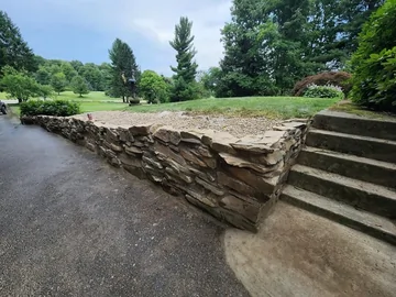 A stone retaining wall separates a gravel area from a paved path and concrete steps, with lush green trees and grass in the background under a partly cloudy sky.