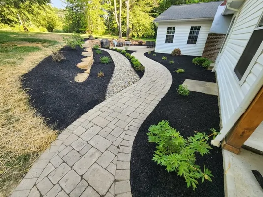 Curved stone walkway bordered by black mulch and green shrubs leads to a patio with patio furniture, next to a white house with brick accents and multiple windows, surrounded by grass and trees.