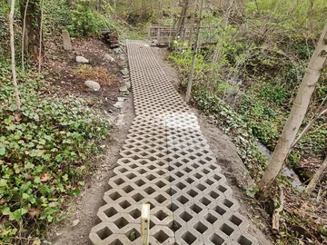 A path made of interlocking concrete bricks with diamond-shaped holes runs through a wooded area with green plants and trees on both sides. A wooden footbridge is visible in the background.