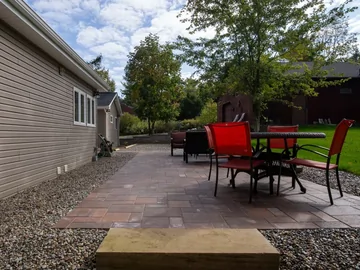 A patio with red and black chairs around a table sits next to a beige house, surrounded by gravel and trees, with green grass and another building in the background under a partly cloudy sky.