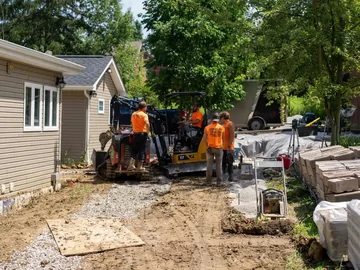 Four construction workers in orange shirts operate machinery and work on a gravel driveway beside a house, surrounded by stacks of materials and trees on a sunny day.
